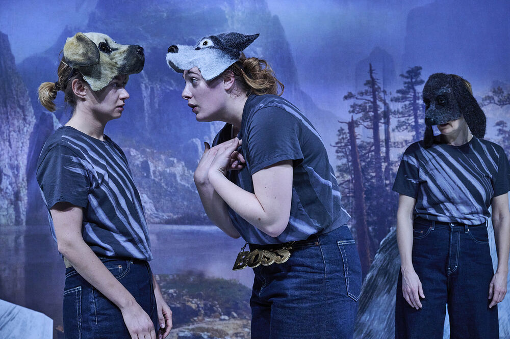 Drei Schauspielerinnen mit Hundemasken. Eine steht abseits, während die anderen beiden die Rangordnung diskutieren. 