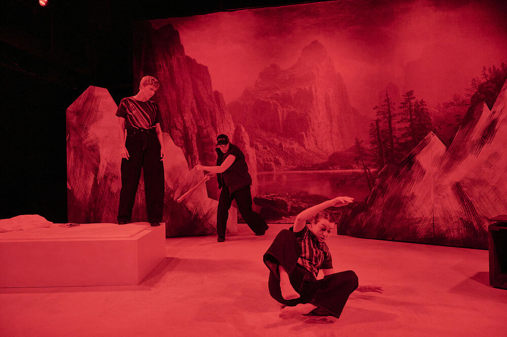 Drei Schauspielerinnen vor einer in rotes Licht getauchten Berglandschaft. Eine steht, eine hält einen Baseballschläger und eine rutscht über den Boden. 